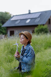 Little girl holding model of wind turbine, standing in front of their house with photovoltaic panels, concept of renewable resources. - HPIF33401