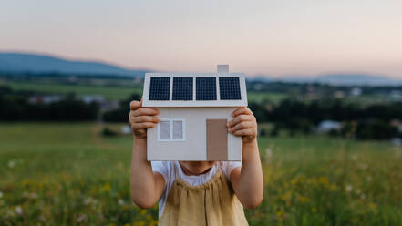 Little girl with model house with installed of solar panels, standing in the middle of meadow. Concept of alternative energy, saving resources and sustainable lifestyle concept. . Importance of alternative energy sources and long-term sustainability for future generations - HPIF33430