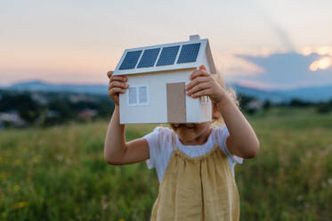 Little girl with model house with installed of solar panels, standing in the middle of meadow. Concept of alternative energy, saving resources and sustainable lifestyle concept. . Importance of alternative energy sources and long-term sustainability for future generations - HPIF33431