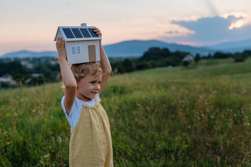Little girl with model house with installed of solar panels, standing in the middle of meadow. Concept of alternative energy, saving resources and sustainable lifestyle concept. . Importance of alternative energy sources and long-term sustainability for future generations - HPIF33433