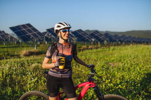 Porträt einer schönen Radfahrerin, die vor den Solarzellen eines Solarparks steht und Wasser trinkt. Ein Solarpark als Lösung für eine nachhaltigere Energiezukunft. - HPIF33436