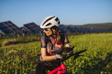 Porträt einer schönen Radfahrerin, die während einer sommerlichen Radtour in der Natur vor den Solarmodulen eines Solarparks steht. Ein Solarpark als Lösung für eine nachhaltigere Energiezukunft. - HPIF33437