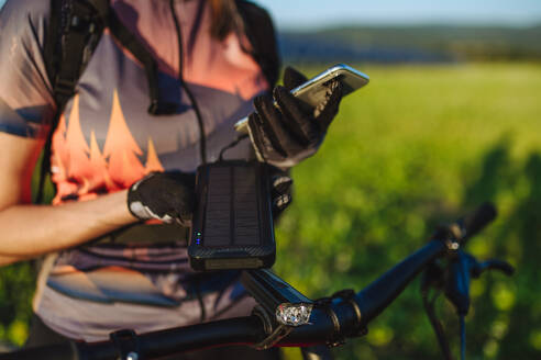 Radfahrerin, die ihr Smartphone mit einem Solarladegerät auflädt. Radfahrerin, die während einer sommerlichen Radtour in der Natur vor den Solarzellen eines Solarparks steht. Konzept der nachhaltigen Energiezukunft. - HPIF33442