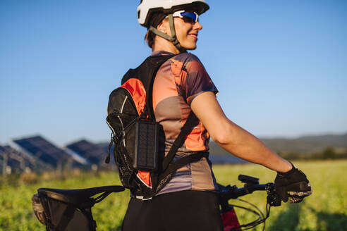 Porträt einer schönen Radfahrerin, die während einer sommerlichen Radtour in der Natur vor den Solarmodulen eines Solarparks steht. Ein Solarpark als Lösung für eine nachhaltigere Energiezukunft. - HPIF33443