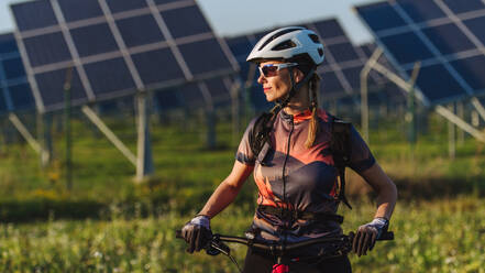 Porträt einer schönen Radfahrerin, die während einer sommerlichen Radtour in der Natur vor den Solarmodulen eines Solarparks steht. Ein Solarpark als Lösung für eine nachhaltigere Energiezukunft. - HPIF33444