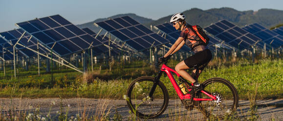 Seitenansicht einer schönen Radfahrerin, die während einer sommerlichen Radtour in der Natur vor den Solarmodulen eines Solarparks fährt. Ein Solarpark als Lösung für eine nachhaltigere Energiezukunft. Sport, Ökologie, Natur-Banner mit Kopierbereich. - HPIF33445