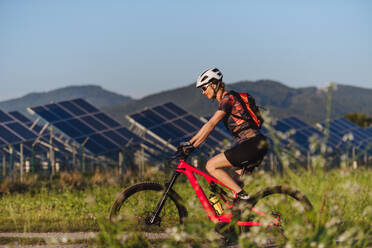 Seitenansicht einer schönen Radfahrerin, die während einer sommerlichen Radtour in der Natur vor den Solarmodulen eines Solarparks fährt. Ein Solarpark als Lösung für eine nachhaltigere Energiezukunft. - HPIF33446