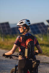 Rückansicht einer schönen Radfahrerin, die während einer sommerlichen Radtour in der Natur vor den Solarmodulen eines Solarparks steht. Ein Solarpark als Lösung für eine nachhaltigere Energiezukunft. - HPIF33449