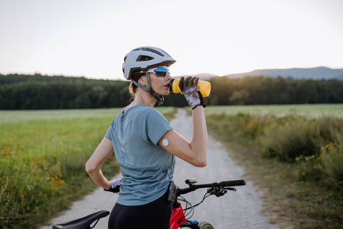 Diabetische Radfahrerin mit einem kontinuierlichen Blutzuckermessgerät am Arm, die während ihrer Radtour Wasser trinkt, um ihren Diabetes während des Trainings in den Griff zu bekommen. Konzept von Bewegung und Diabetes. - HPIF33450