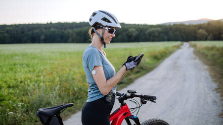 Diabetische Radfahrerin mit kontinuierlichem Blutzuckermessgerät, die ihre Blutzuckerwerte in Echtzeit auf dem Smartphone kontrolliert. Konzept von Bewegung und Diabetes. - HPIF33453