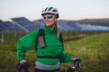 Porträt einer schönen Radfahrerin, die während einer sommerlichen Radtour in der Natur vor den Solarmodulen eines Solarparks steht. Ein Solarpark als Lösung für eine nachhaltigere Energiezukunft. - HPIF33459