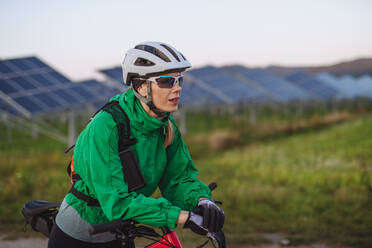 Porträt einer schönen Radfahrerin, die während einer sommerlichen Radtour in der Natur vor den Solarmodulen eines Solarparks steht. Ein Solarpark als Lösung für eine nachhaltigere Energiezukunft. - HPIF33460