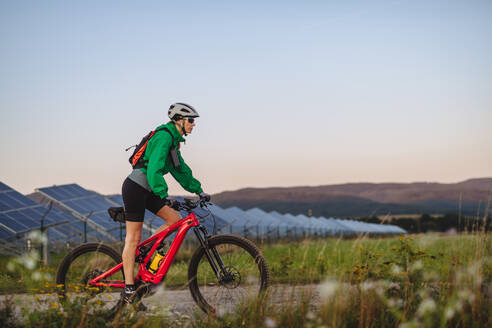 Seitenansicht einer schönen Radfahrerin, die während einer sommerlichen Radtour in der Natur vor den Solarmodulen eines Solarparks fährt. Ein Solarpark als Lösung für eine nachhaltigere Energiezukunft. - HPIF33461