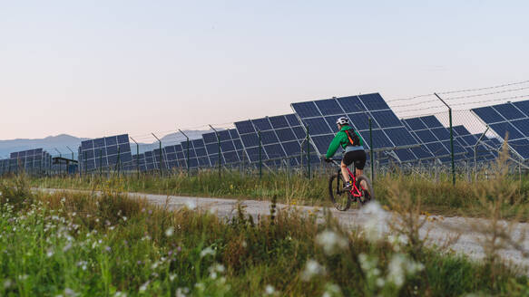 Rückansicht einer Radfahrerin, die während einer sommerlichen Radtour in der Natur vor den Solarmodulen eines Solarparks fährt. Ein Solarpark als Lösung für eine nachhaltigere Energiezukunft. Banner mit Werbefläche. - HPIF33462