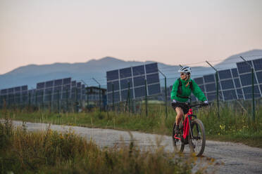 Radfahrerin, die während einer sommerlichen Radtour in der Natur vor den Solarmodulen eines Solarparks fährt. Ein Solarpark als Lösung für eine nachhaltigere Energiezukunft. Banner mit Kopierbereich. - HPIF33463