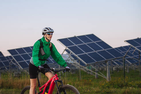 Porträt einer schönen Radfahrerin, die während einer sommerlichen Radtour in der Natur vor den Solarmodulen eines Solarparks fährt. Ein Solarpark als Lösung für eine nachhaltigere Energiezukunft. - HPIF33464