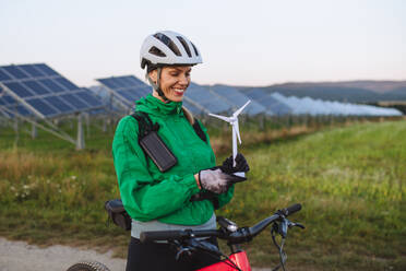 Porträt einer schönen Radfahrerin, die während einer sommerlichen Radtour in der Natur vor Sonnenkollektoren in einem Solarpark steht. Radfahrerin mit Modell einer Windkraftanlage. Alternative und nachhaltige Energiequellen. - HPIF33466