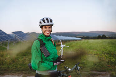 Porträt einer schönen Radfahrerin, die während einer sommerlichen Radtour in der Natur vor Sonnenkollektoren in einem Solarpark steht. Radfahrerin mit Modell einer Windkraftanlage. Alternative und nachhaltige Energiequellen. - HPIF33467