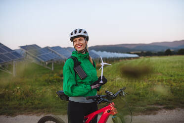 Porträt einer schönen Radfahrerin, die während einer sommerlichen Radtour in der Natur vor Sonnenkollektoren in einem Solarpark steht. Radfahrerin mit Modell einer Windkraftanlage. Alternative und nachhaltige Energiequellen. - HPIF33468