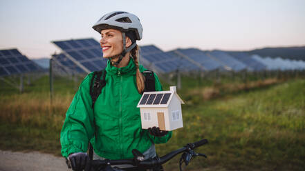 Porträt einer schönen Radfahrerin, die während einer sommerlichen Radtour in der Natur vor Solarmodulen in einem Solarpark steht. Radfahrer hält ein Modell eines Hauses mit Photovoltaik-Modulen auf dem Dach. Solarenergie - nachhaltige Energiezukunft. - HPIF33469