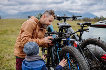 Kleiner Junge hilft seinem Vater, das Fahrrad auf dem Gepäckträger zu befestigen. Vorbereitung für eine Fahrradtour in der Natur, Abstellen der Fahrräder von den Autoträgern. Konzept der gesunden Lebensweise und der Bewegung. - HPIF33475
