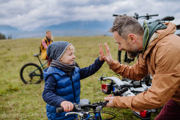 Junge Familie mit kleinen Kindern, die sich auf eine Fahrradtour in der Natur vorbereiten, indem sie die Fahrräder von den Autoträgern abstellen. Vater und Sohn geben sich die Hand und freuen sich auf die gemeinsame Fahrt. Konzept des gesunden Lebensstils und der bewegten Aktivität. - HPIF33478