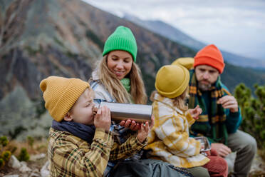 Glückliche Familie, die sich ausruht und einen Snack zu sich nimmt, während sie gemeinsam in den herbstlichen Bergen wandern. Ein kleiner Junge trinkt Tee aus einer Thermoskanne. - HPIF33536