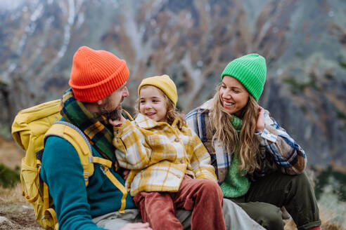 Eine glückliche Familie, die sich ausruht, einen Snack zu sich nimmt und die Aussicht beim gemeinsamen Wandern in den herbstlichen Bergen genießt. Mutter, Vater und Tochter auf einer kinderfreundlichen Wanderung. - HPIF33540