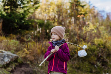 Porträt eines kleinen Mädchens mit einer Windel während einer Herbstwanderung in den Bergen. Das Mädchen hält einen Stock mit einem Tuch in der Hand und trägt einen Snack darin. - HPIF33561