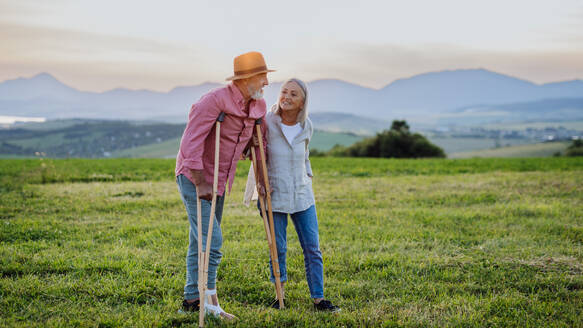 Senior man with broken leg and crutches standing in the the middle of meadow. Recovery, rehabilitation after injury or surgery in the nature. Wife helping husband with walking during easy walk on their vacation. - HPIF33596