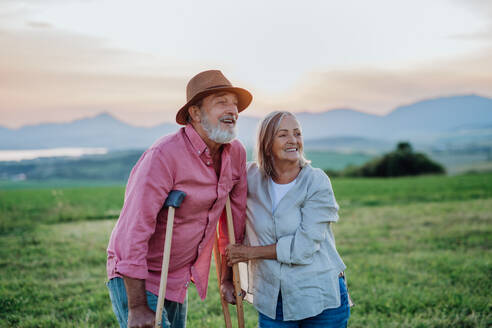 Senior man with broken leg and crutches standing in the the middle of meadow. Recovery, rehabilitation after injury or surgery in the nature. Wife helping husband with walking during easy walk on their vacation. Concept of traveling with physical disabilities. - HPIF33597