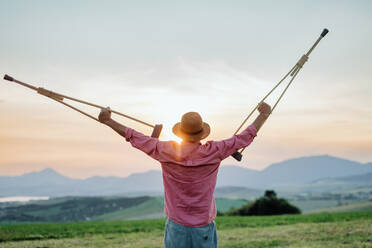 Rear view of senior man holding crutches in the air, he is happy abtou his recovery. Recovery, rehabilitation after injury or surgery in the nature. Concept of recovery and physical therapy - HPIF33599