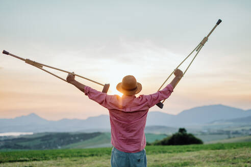 Rear view of senior man holding crutches in the air, he is happy abtou his recovery. Recovery, rehabilitation after injury or surgery in the nature. Concept of recovery and physical therapy - HPIF33599