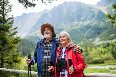 Active elderly couple hiking together in autumn mountains, on senior friendly trail. Senior spouses on the vacation in the mountains celebrating anniversary. Senior tourists with backpacks using trekking poles for stability. - HPIF33607
