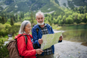 Active elderly couple hiking together in autumn mountains, on senior friendly trail. Senior spouses on the vacation in the mountains celebrating anniversary. Senior tourists with backpacks reading map, preparing for hike. - HPIF33611