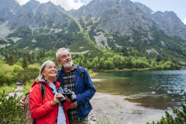 Potrait einer aktiven älteren Frau, die durch ein Fernglas in die Natur blickt. Älteres Paar beim Wandern in den herbstlichen Bergen auf einem seniorenfreundlichen Wanderweg. Älterer Tourist mit Rucksack, der Trekkingstöcke zur Stabilität benutzt. - HPIF33618