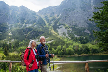 Active elderly couple hiking together in autumn mountains, on senior-friendly trail. Husband and wife walking by lake, enjoying nature. Senior tourists using trekking poles for stability. - HPIF33620