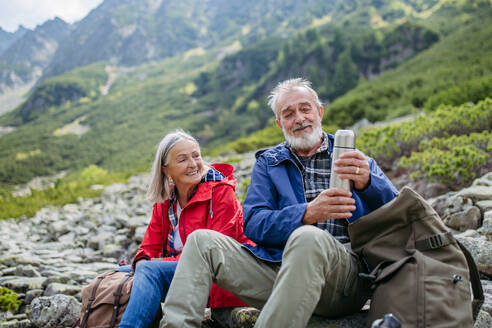 Aktives älteres Ehepaar beim Wandern in den herbstlichen Bergen, auf einem seniorenfreundlichen Wanderweg. Mann und Frau trinken warmen Tee, Kaffee aus einer Thermoskanne. Seniorentourist mit Rucksack, der sich während der Wanderung ausruht. - HPIF33625