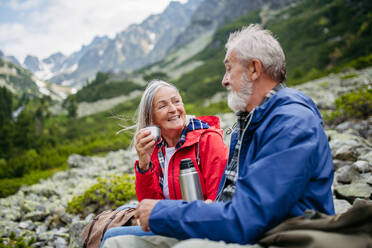 Aktives älteres Ehepaar beim Wandern in den herbstlichen Bergen, auf einem seniorenfreundlichen Wanderweg. Mann und Frau trinken warmen Tee, Kaffee aus einer Thermoskanne. Seniorentourist mit Rucksack, der sich während der Wanderung ausruht. - HPIF33628