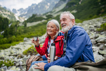 Aktives älteres Ehepaar beim Wandern in den herbstlichen Bergen, auf einem seniorenfreundlichen Wanderweg. Mann und Frau trinken warmen Tee, Kaffee aus einer Thermoskanne. Seniorentourist mit Rucksack, der sich während der Wanderung ausruht. - HPIF33629