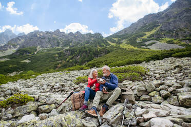 Aktives älteres Paar beim Wandern in den herbstlichen Bergen, auf einem seniorenfreundlichen Wanderweg. Mann und Frau nehmen einen gesunden Snack zu sich, um Energie zu tanken. Ältere Touristen mit Rucksäcken ruhen sich beim Wandern aus. - HPIF33633