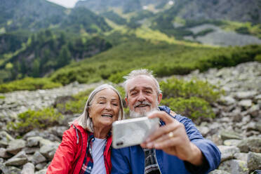 Active elderly couple hiking together in autumn mountains, on senior friendly trail. Husband and wife taking selfie with smartphone. Senior tourist with backpacks getting rest during hike. - HPIF33635