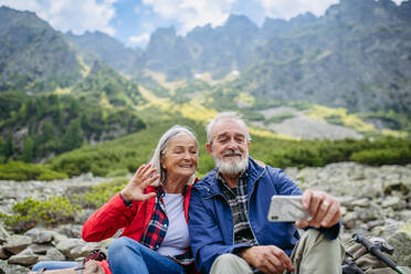 Active elderly couple hiking together in autumn mountains, on senior friendly trail. Husband and wife taking selfie with smartphone. Senior tourist with backpacks getting rest during hike. - HPIF33636