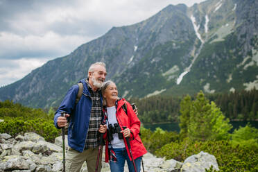 Waist up shot of active elderly couple hiking together in autumn mountains, on senior friendly trail. Husband and wife getting rest during hike, enjoying nature. Senior tourist with backpack using trekking poles for stability. - HPIF33642