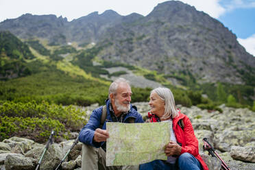 Active elderly couple hiking together in autumn mountains, on senior friendly trail. Senior spouses on the vacation in the mountains celebrating anniversary. Senior tourists sitting on rocks and looking at the route on a map. - HPIF33644