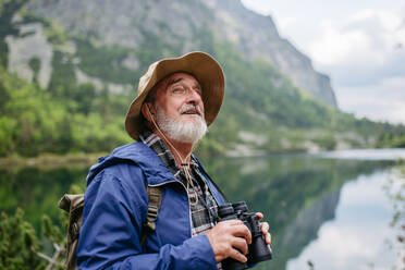 Potrait eines aktiven älteren Mannes beim Wandern in den herbstlichen Bergen, auf einem seniorenfreundlichen Wanderweg. Ein älterer Tourist mit Rucksack schaut durch ein Fernglas. - HPIF33650