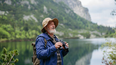 Potrait eines aktiven älteren Mannes beim Wandern in den herbstlichen Bergen, auf einem seniorenfreundlichen Wanderweg. Ein älterer Tourist mit Rucksack schaut durch ein Fernglas. - HPIF33651