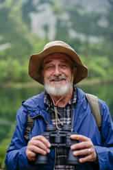 Potrait eines aktiven älteren Mannes beim Wandern in den herbstlichen Bergen, auf einem seniorenfreundlichen Wanderweg. Ein älterer Tourist mit Rucksack schaut durch ein Fernglas. - HPIF33652