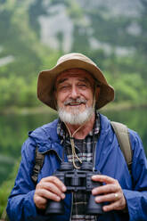 Potrait of active senior man hiking in autumn mountains, on senior friendly trail. Senior tourist with backpack looking through binoculars. - HPIF33652