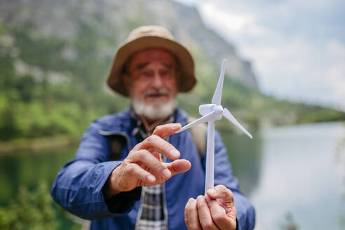 Potrait eines aktiven älteren Touristen, der ein Modell einer Windturbine hält. Ein älterer Mann mit Rucksack, der die Flügel der Turbine dreht. Konzept der erneuerbaren, grünen Energie und des nachhaltigen Lebensstils. - HPIF33656